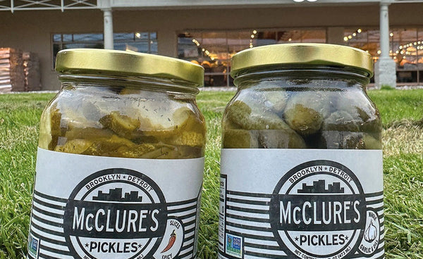 Two jars of McClure's Pickles on grass with a building in the background